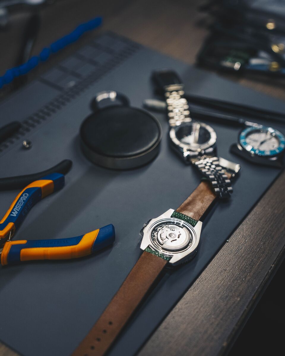 A bench flat lay of pliers, a case puller, a bracelet and a watch on a tan strap showing its caseback.
